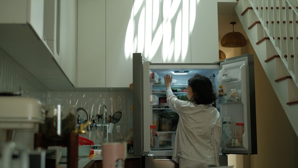 Person im Schlafanzug steht vor dem geöffneten Kühlschrank in der Küche